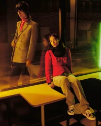 Two fashion models posing near a window booth in warm cinematic lighting with neon green accents.