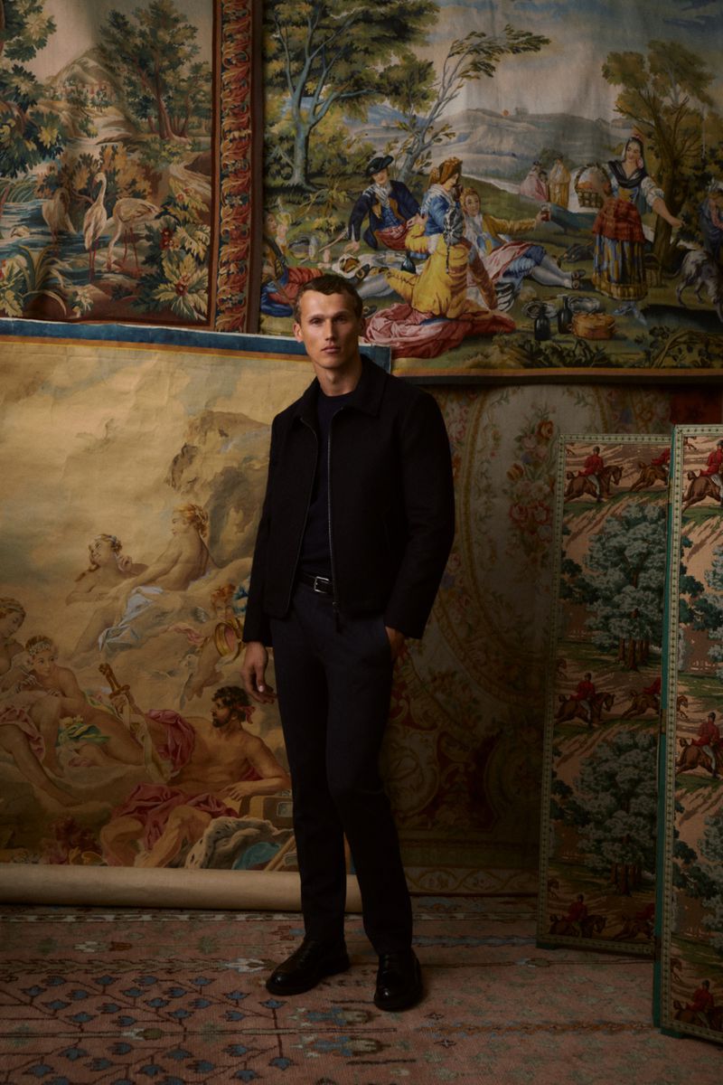 Man in a black jacket and dark trousers stands in front of tapestry art in an indoor setting. Fashion shot, full body.