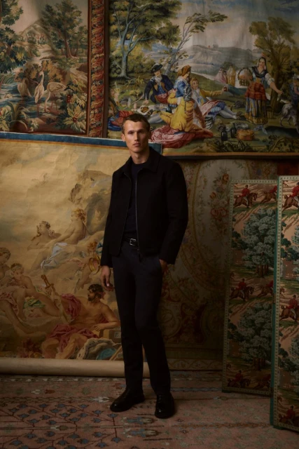 Man in a black jacket and dark trousers stands in front of tapestry art in an indoor setting. Fashion shot, full body.