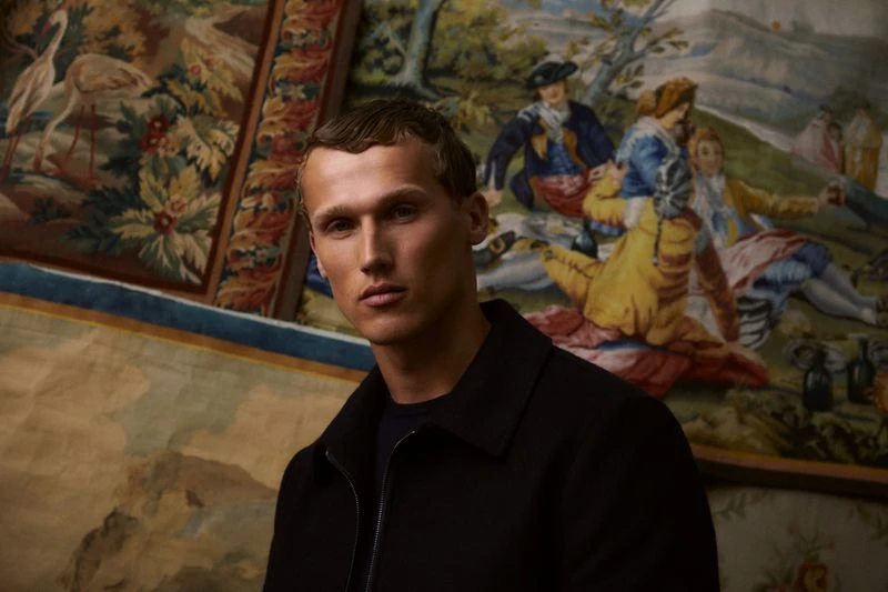 Man in a dark jacket poses indoors with a tapestry backdrop; neutral expression and soft lighting create a sophisticated portrait.