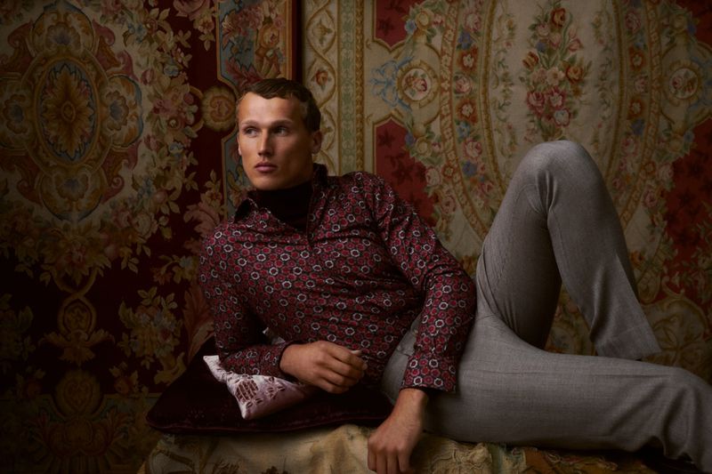 Man in patterned shirt and grey pants lying against antique patterned rug and pillow, looking away in a fashion editorial setting.