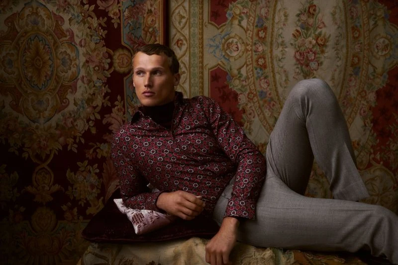 Man in patterned shirt and grey pants lying against antique patterned rug and pillow, looking away in a fashion editorial setting.