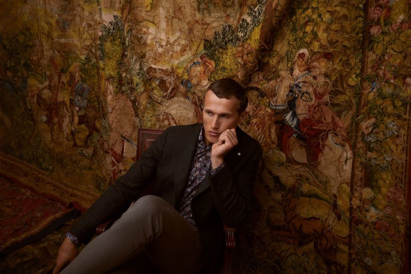Man in blazer and light-grey pants sitting on a vintage chair in an indoor setting with a tapestry behind him, looking off-camera.