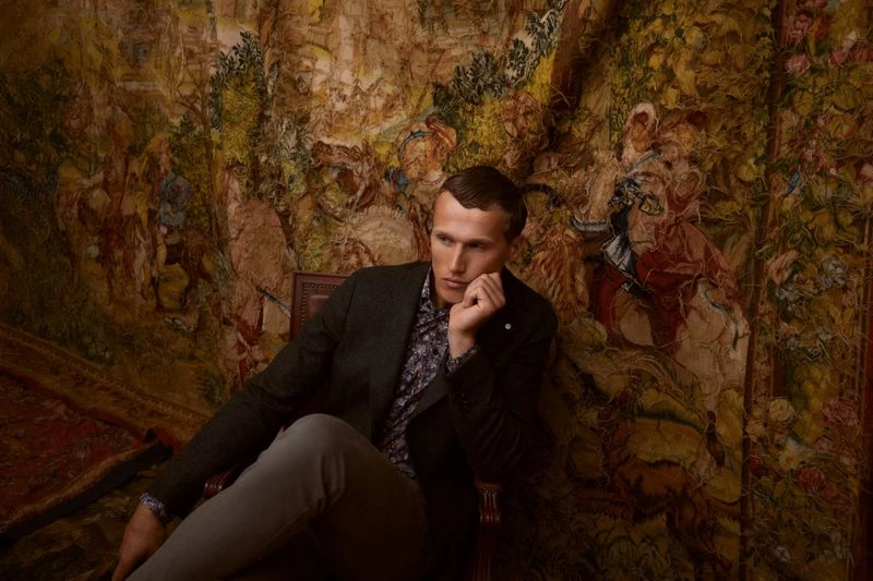 Man in blazer and light-grey pants sitting on a vintage chair in an indoor setting with a tapestry behind him, looking off-camera.