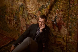 A man in a dark blazer and patterned shirt seated against an ornate antique tapestry background for the MIRTO Campaign Man FW25.
