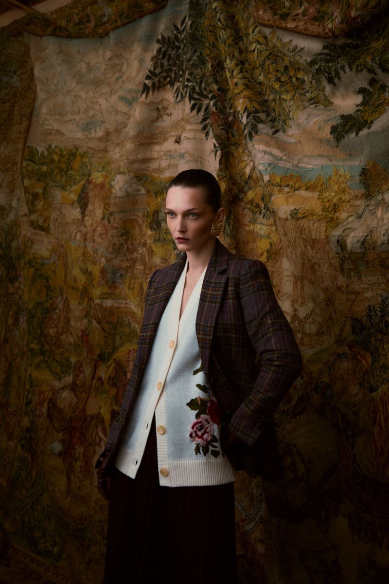 Woman in a plaid blazer and floral cardigan poses against a painted backdrop, fashion portrait, looking serious, indoor setting.