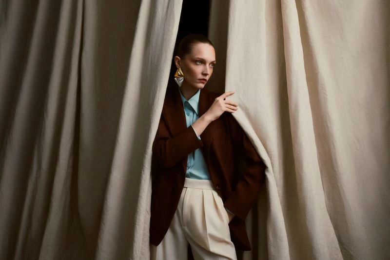 Woman posing in brown jacket and white pants, standing between beige curtains, with golden earrings in a studio fashion shot.