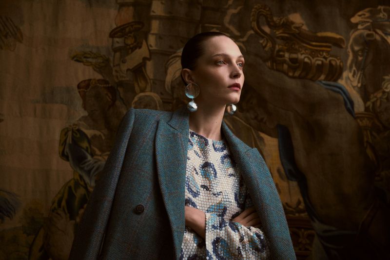 Elegant woman in a teal coat and patterned dress stands indoors against a backdrop of a classic mural, showcasing fashion and style.