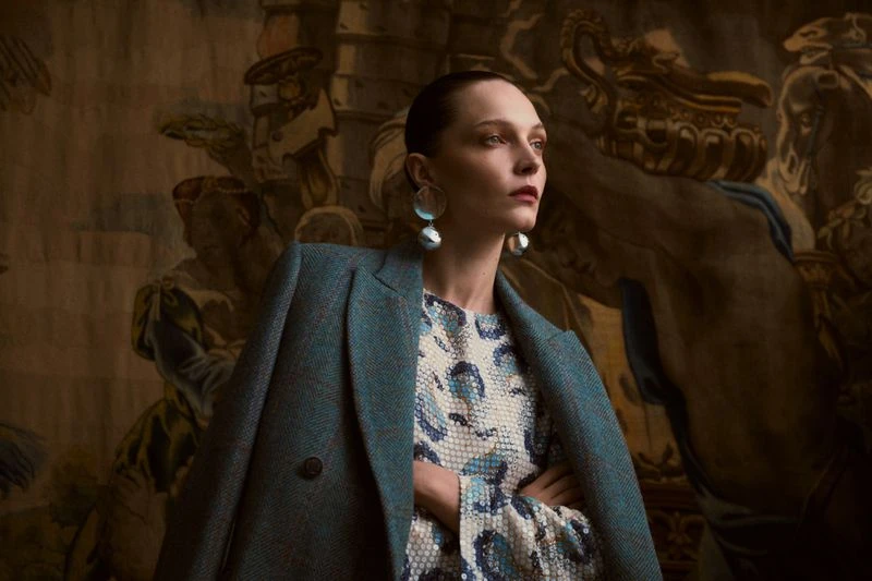 Elegant woman in a teal coat and patterned dress stands indoors against a backdrop of a classic mural, showcasing fashion and style.