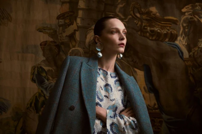Elegant woman in a teal coat and patterned dress stands indoors against a backdrop of a classic mural, showcasing fashion and style.