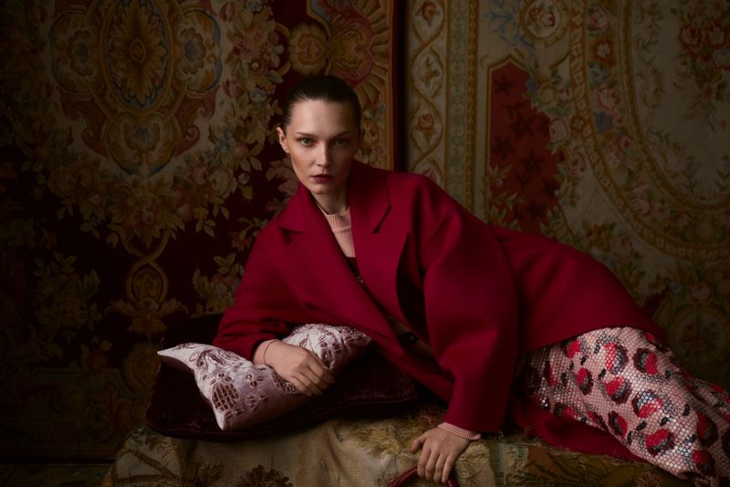 A woman in a red coat lies on a patterned surface with an ornate floral textile backdrop, creating a rich, luxurious fashion image.