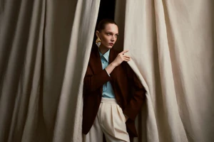 Model wearing a brown blazer, light blue shirt, and cream pleated trousers looking sideways from behind draped beige curtains.
