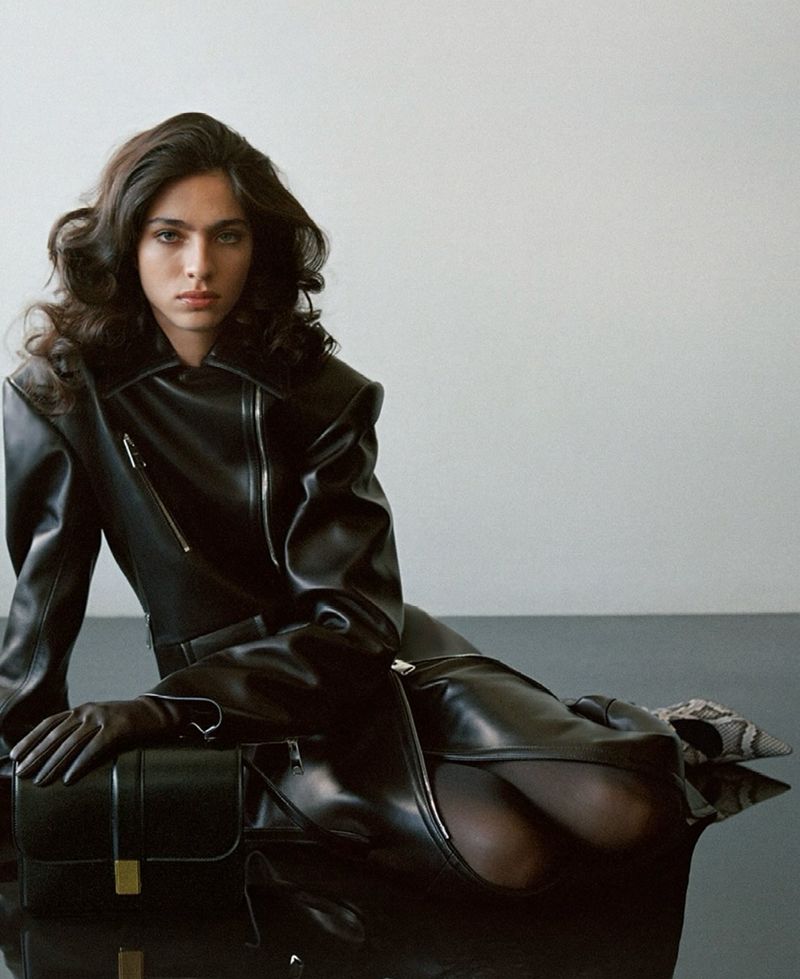 Adult female model poses in shiny black leather coat and gloves next to black handbag against a neutral studio backdrop.