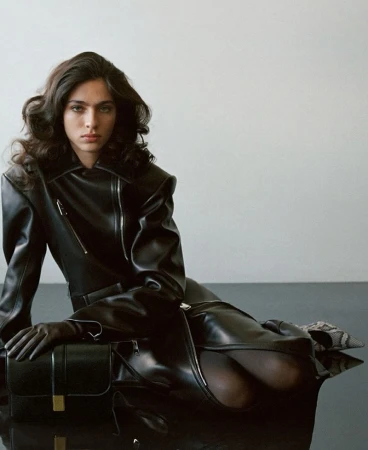 Model Loli Bahia seated in a black leather jacket dress with leather gloves, black handbag, and snakeskin pumps.