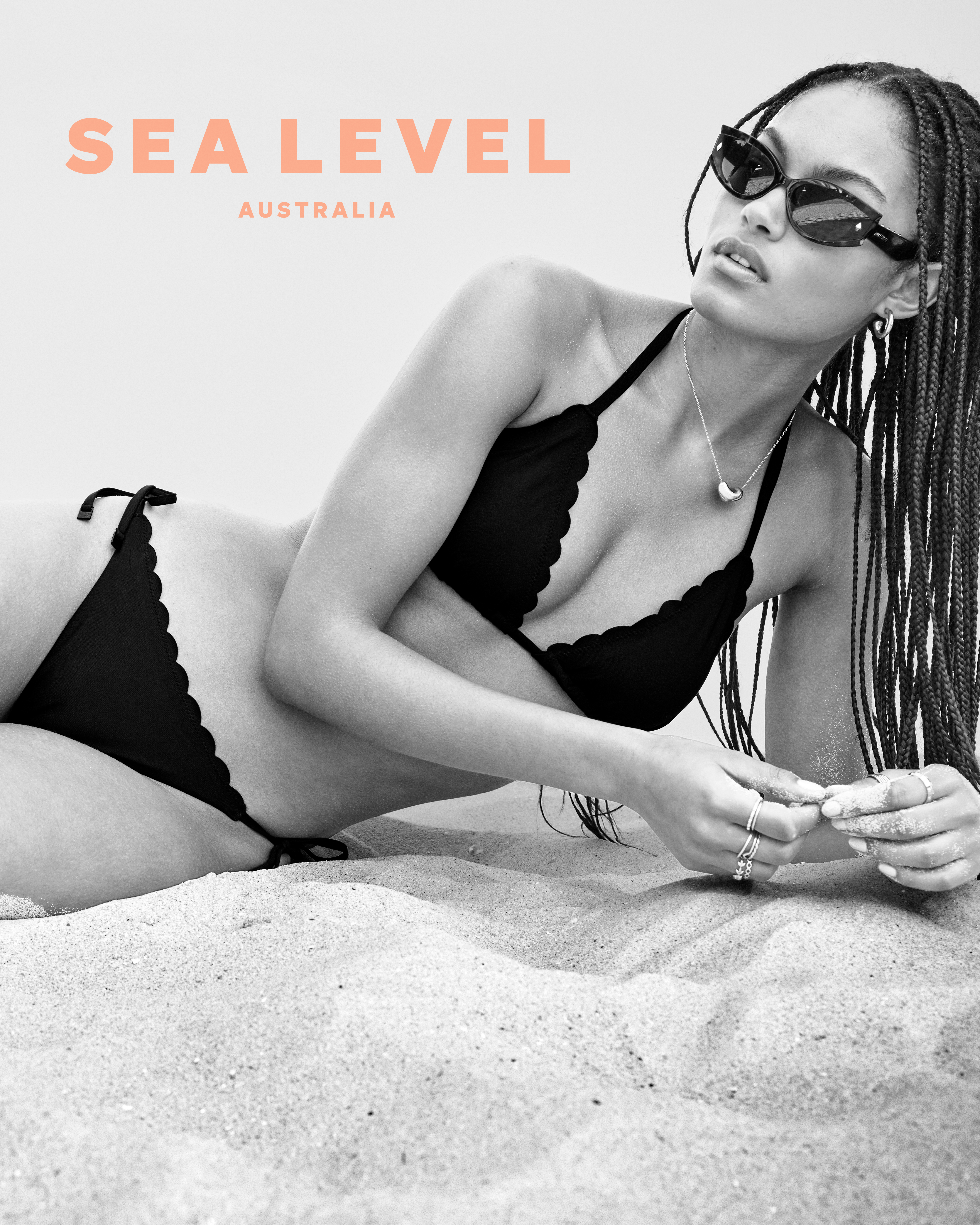 Woman in black bikini and sunglasses lying on sand, advertising Sea Level Australia swimwear, summer fashion, beach lifestyle.
