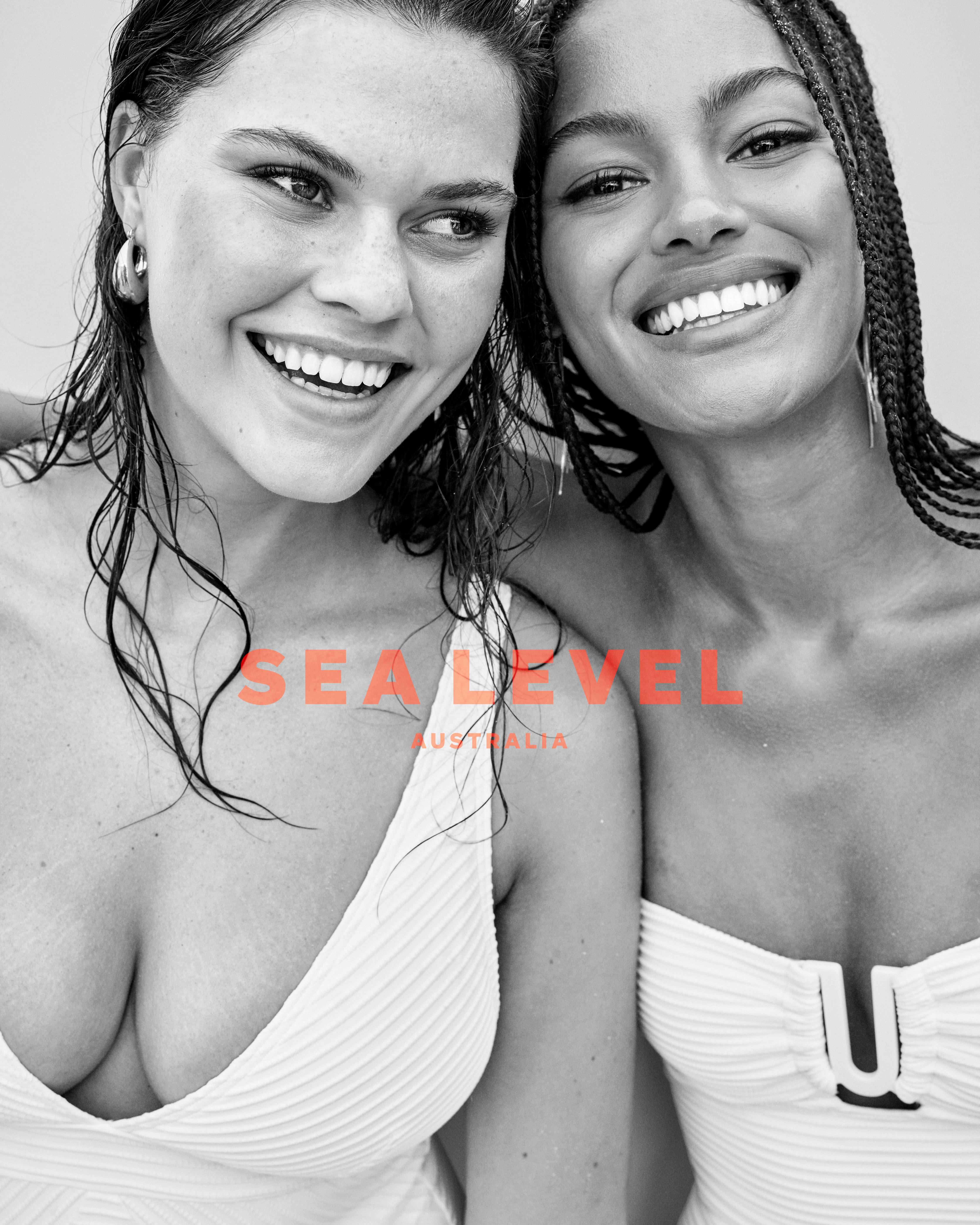Two women smile in white swimsuits in a bright studio shot featuring the Sea Level Australia logo and a monochromatic color scheme.