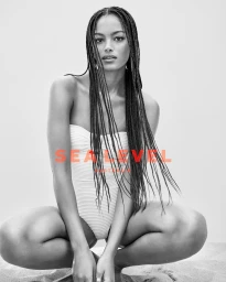 Model with long braided hair wearing a white strapless swimsuit crouching outdoors