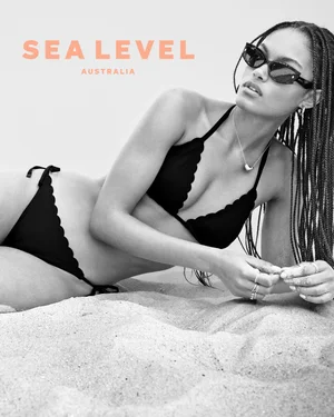 Model in black bikini and sunglasses lying on a sandy beach for Sea Level Australia