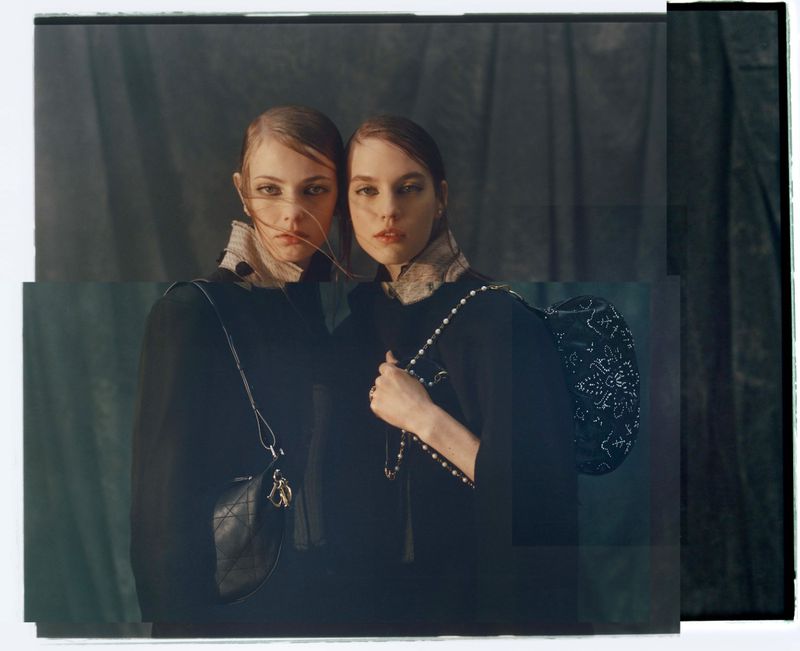 Two fashion models in dark coats and handbags pose indoors against a draped dark background. One woman has a pearl-beaded bag.
