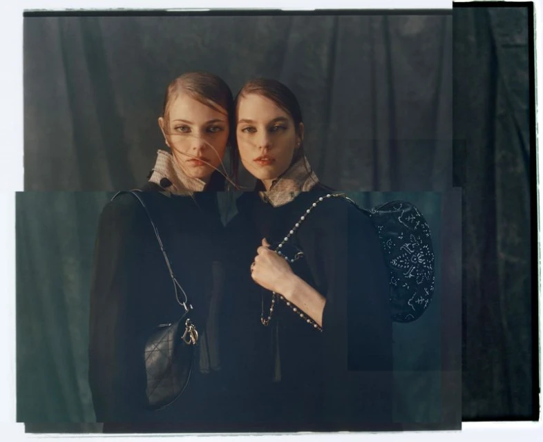 Two fashion models in dark coats and handbags pose indoors against a draped dark background. One woman has a pearl-beaded bag.