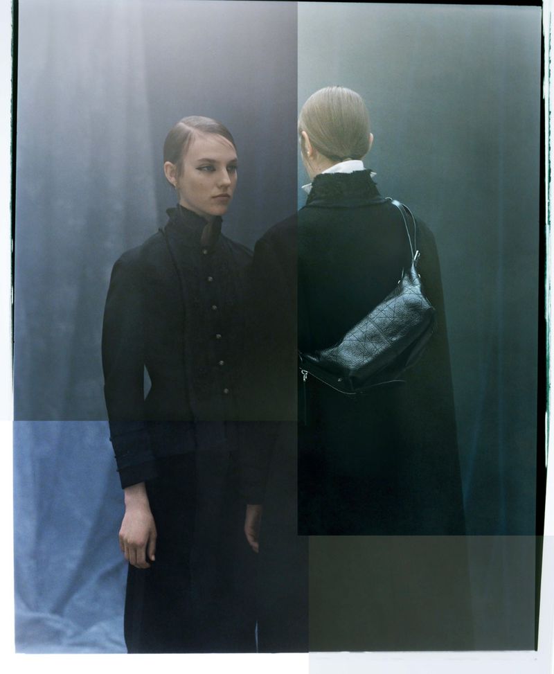 Two women in dark coats, one facing forward with a shoulder bag, in a soft, desaturated studio setting with moody lighting and cool tones.