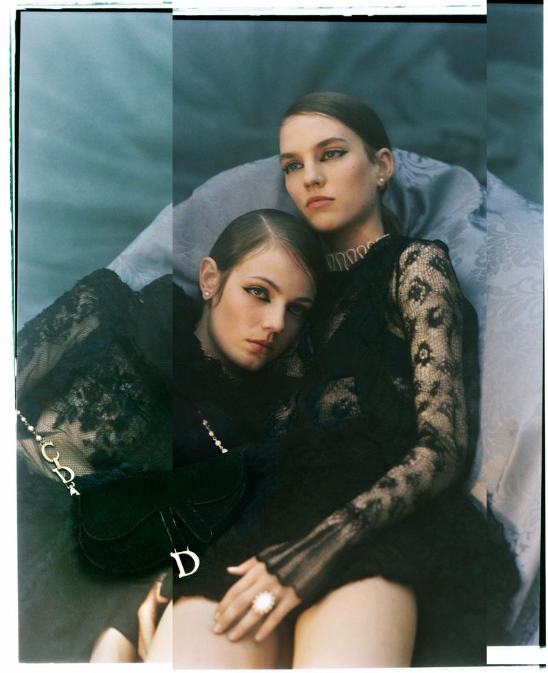 Two young women pose intimately in a studio wearing black lace tops, complemented by a black purse. Soft, diffused lighting enhances the mood.