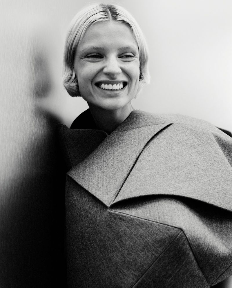 Black and white portrait of a smiling blonde woman with short hair wearing a gray, geometrically designed top in front of a light wall.