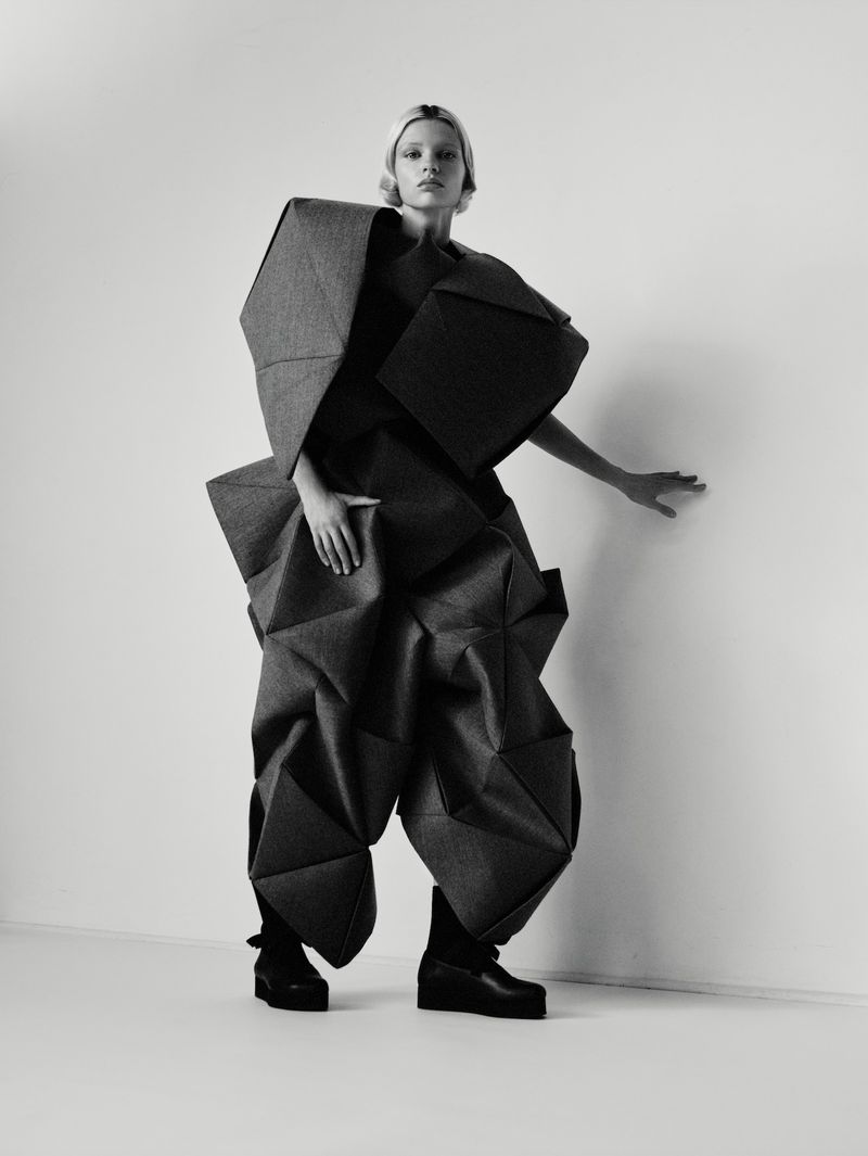 Monochrome fashion shot of a fair-skinned model wearing a grey, geometrically designed outfit in a photo studio setting.