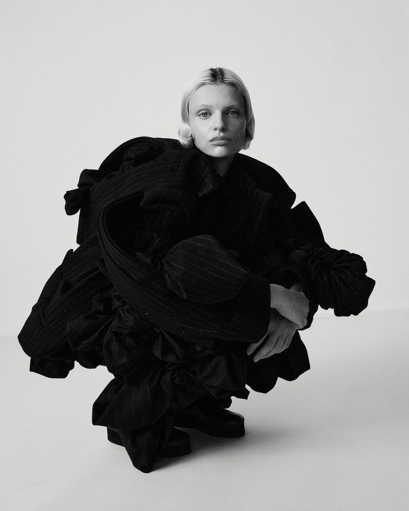 Monochrome fashion shot of an adult female crouched in a photo studio, wearing an oversized pinstripe jacket and dark footwear.
