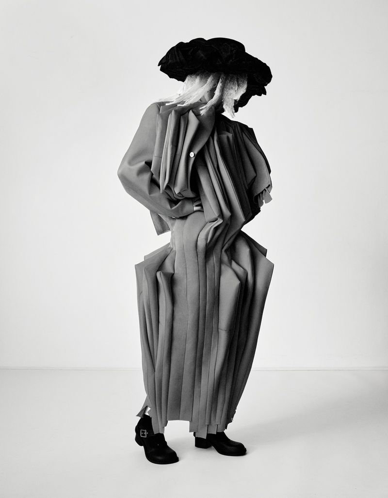 Fashionable individual in an avant-garde gray outfit and oversized black hat strikes a pose in a clean photo studio setting.