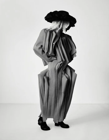 Fashionable individual in an avant-garde gray outfit and oversized black hat strikes a pose in a clean photo studio setting.