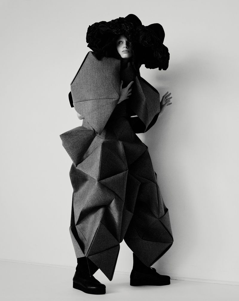 Fashion model poses in a grey geometric garment with large black headdress against a plain white studio backdrop for a high fashion editorial.