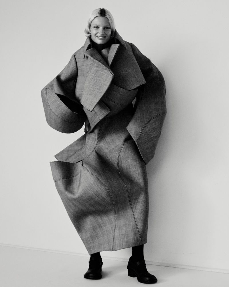 A smiling woman with platinum blonde hair poses indoors wearing a futuristic gray coat and skirt with black boots against a white wall.