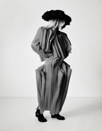 Model Mila van Eeten posing in a sculptural pleated tailored coat and oversized black hat against a plain light studio backdrop.
