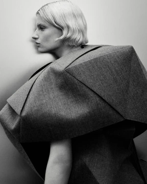 Model in a structured grey sculptural garment against a plain backdrop in a black and white fashion portrait.