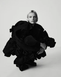 Model wearing voluminous dark layered jacket crouches against a plain light studio background for Purple Magazine.