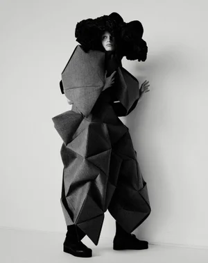 Model Mila van Eeten wearing an avant-garde geometric folded garment and black headpiece against a plain studio background.