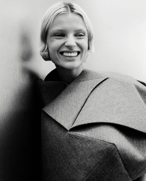 Model Mila van Eeten smiling while wearing a sculptural geometric grey coat against a smooth backdrop.