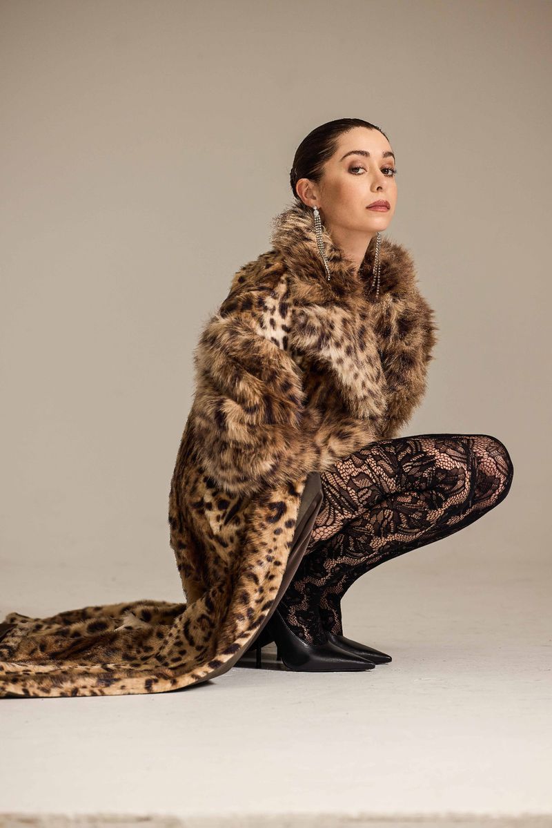 Woman in faux fur leopard coat and black lace tights in a studio portrait, kneeling on the floor and looking at the camera.