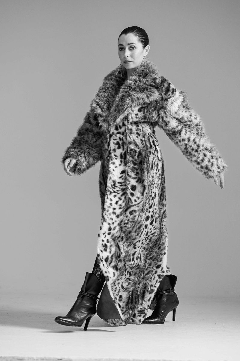 A woman in a faux fur coat with an animal print, black leggings, and black heeled boots stands in a photo studio with neutral background.