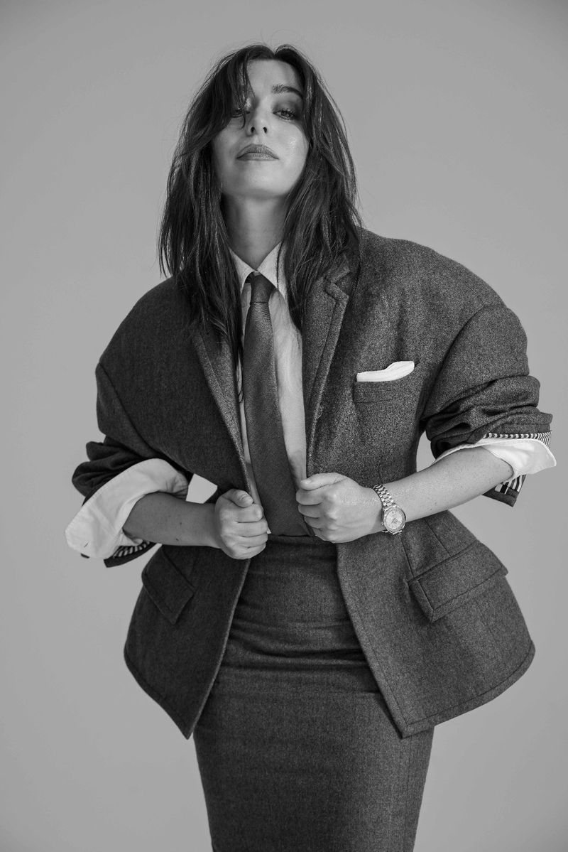 A stylish woman in a gray skirt suit and tie poses confidently against a neutral backdrop, creating a bold and sophisticated visual.