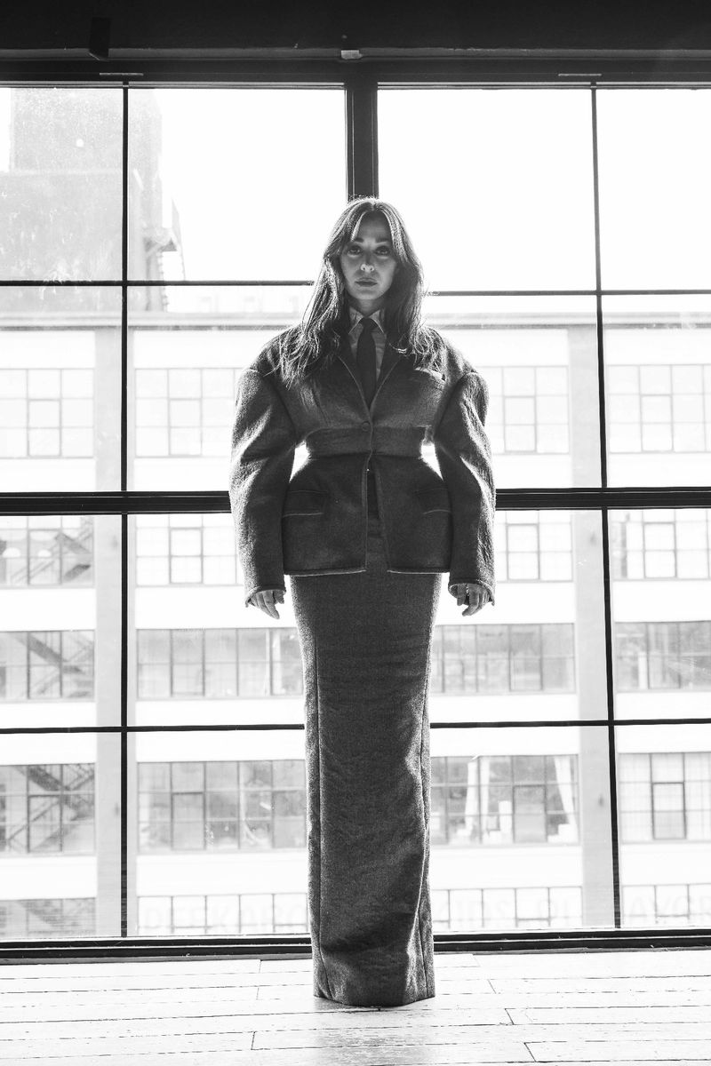 Monochrome portrait of a woman in a jacket and long skirt, posing in front of a window with city buildings in the background.