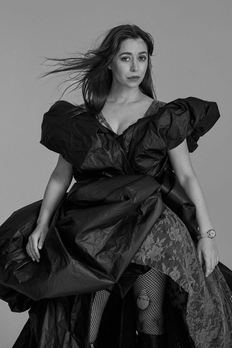 Monochrome portrait of a woman in a sculptural black dress with lace details and fishnet stockings against a grey backdrop, dramatic lighting.
