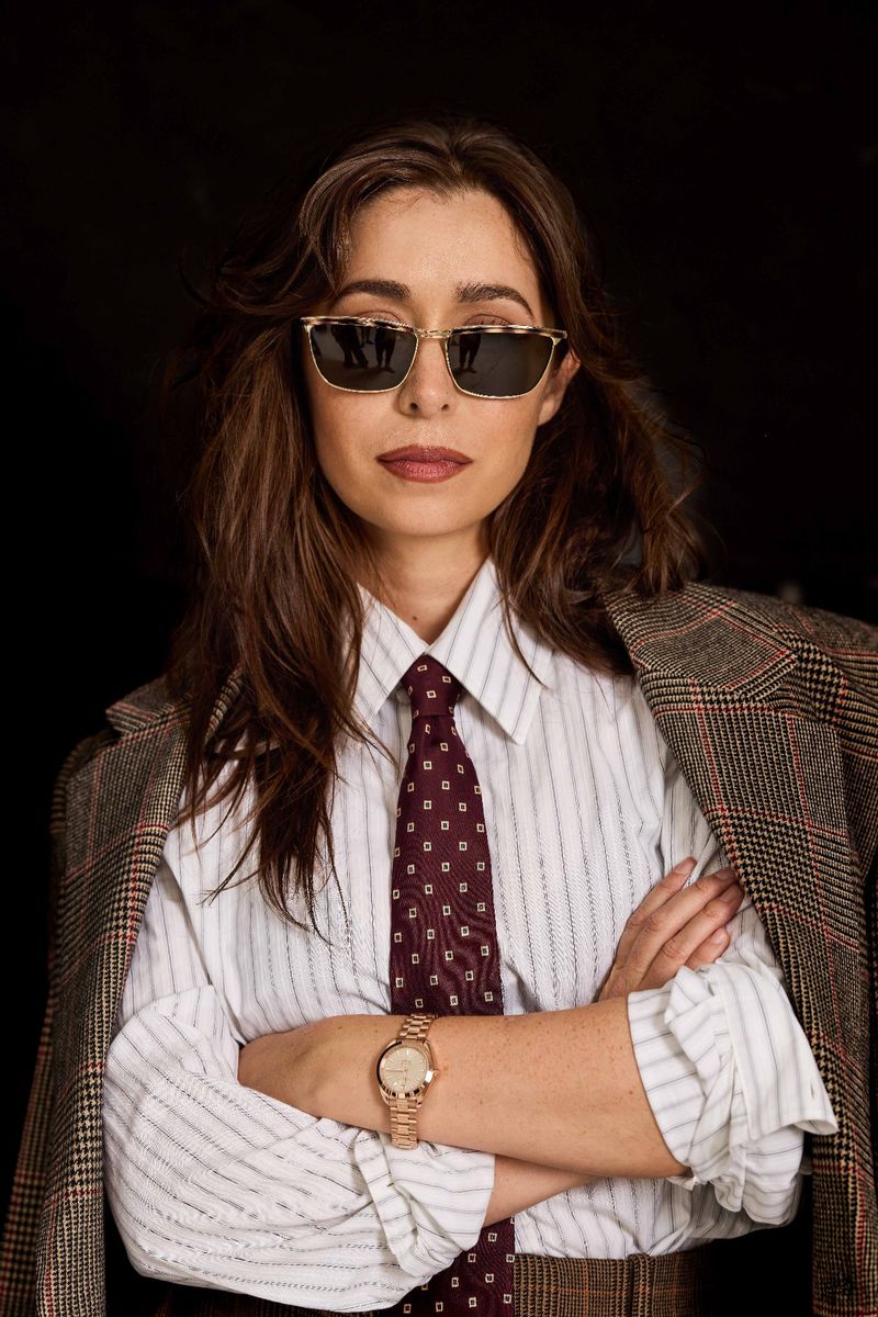 Stylish woman in striped shirt, tie, sunglasses and blazer against a dark background, posing with arms crossed, looking at the viewer.