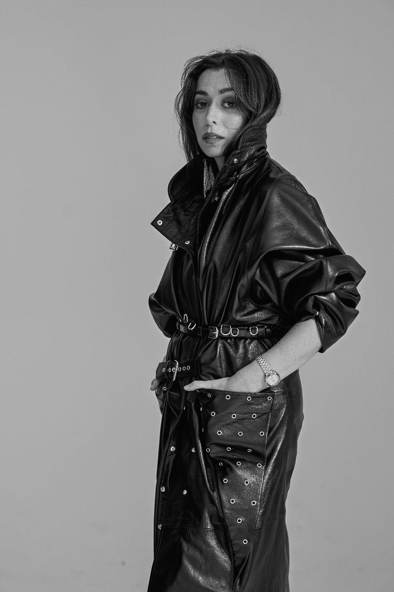 Black and white portrait of adult female in long black leather coat with a belt, standing indoors with a neutral backdrop.