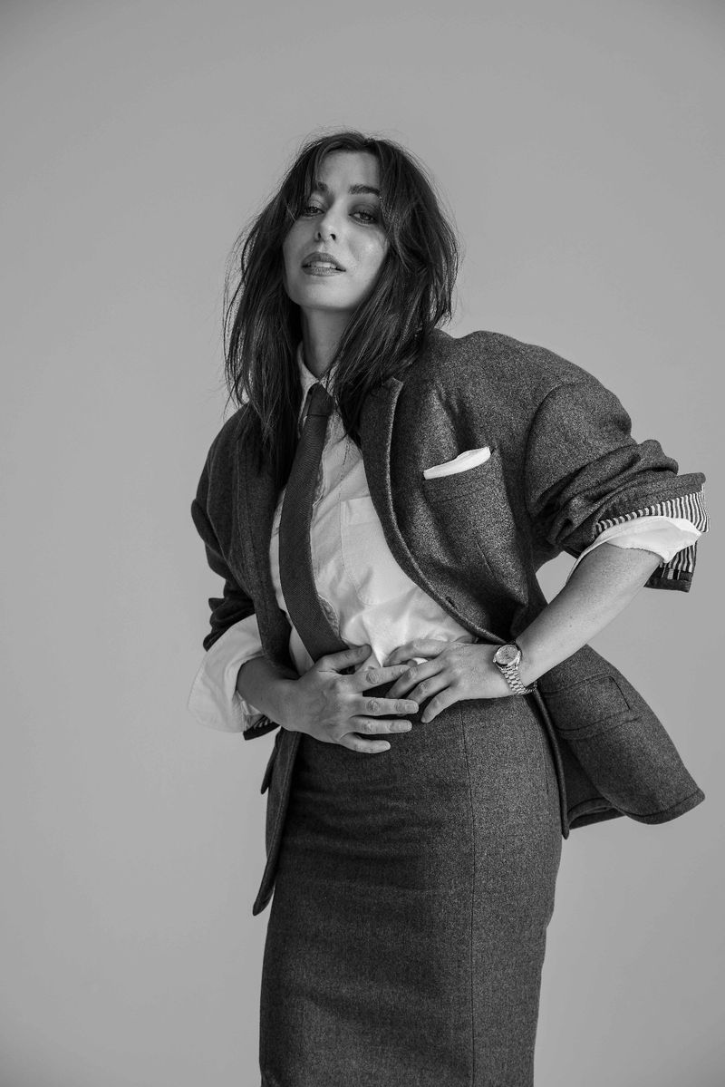 Black and white portrait of a woman in a tailored skirt suit, white shirt, and tie, posing in a photo studio setting with neutral background.