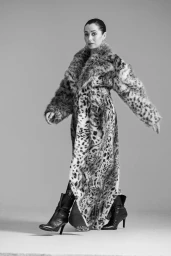 Cristin Milioti wearing an oversized leopard print fur coat and black heeled boots against a grey studio backdrop.