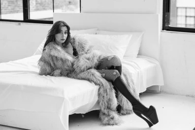 Cristin Milioti wearing a shaggy faux fur coat, ripped fishnets, and platform boots while reclining on a white bed in black and white.