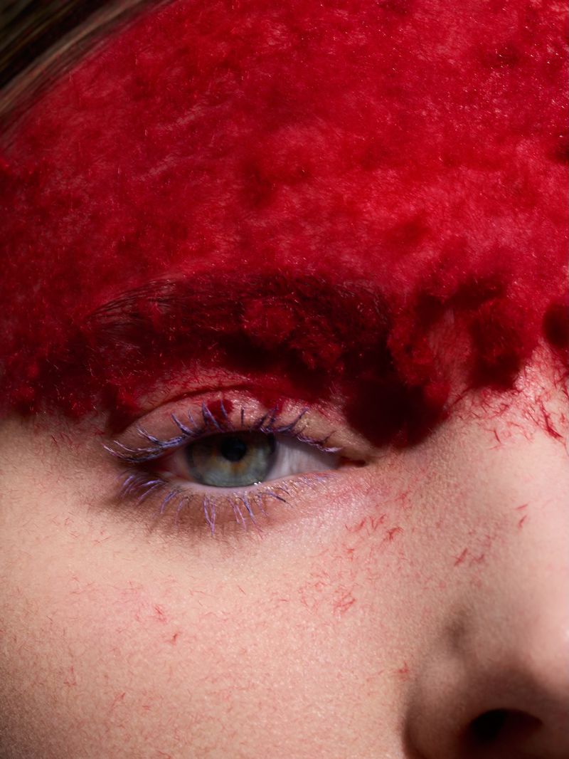 Macro shot of a person's face with vivid red dye covering the upper forehead and bright blue mascara on the eyelashes.