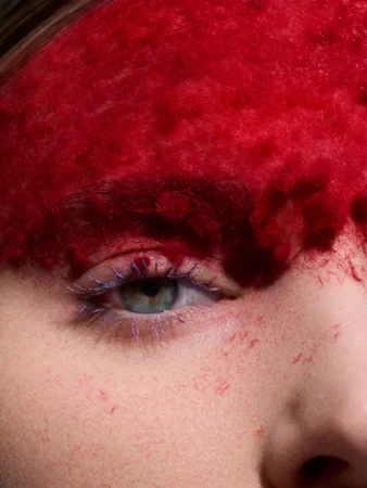 Macro shot of a person's face with vivid red dye covering the upper forehead and bright blue mascara on the eyelashes.